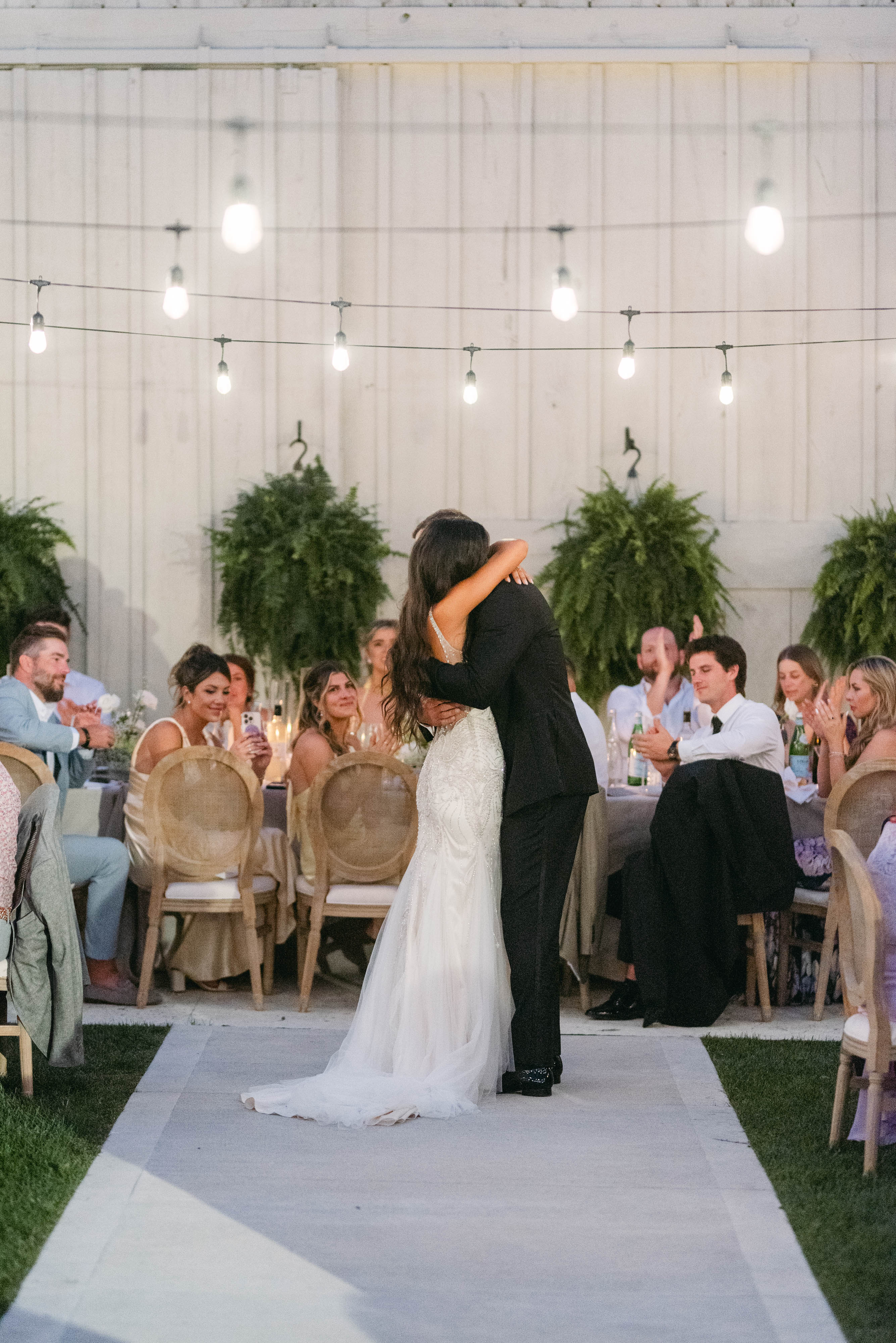Bride and groom's first dance embrace under string lights and hanging ferns at the barn