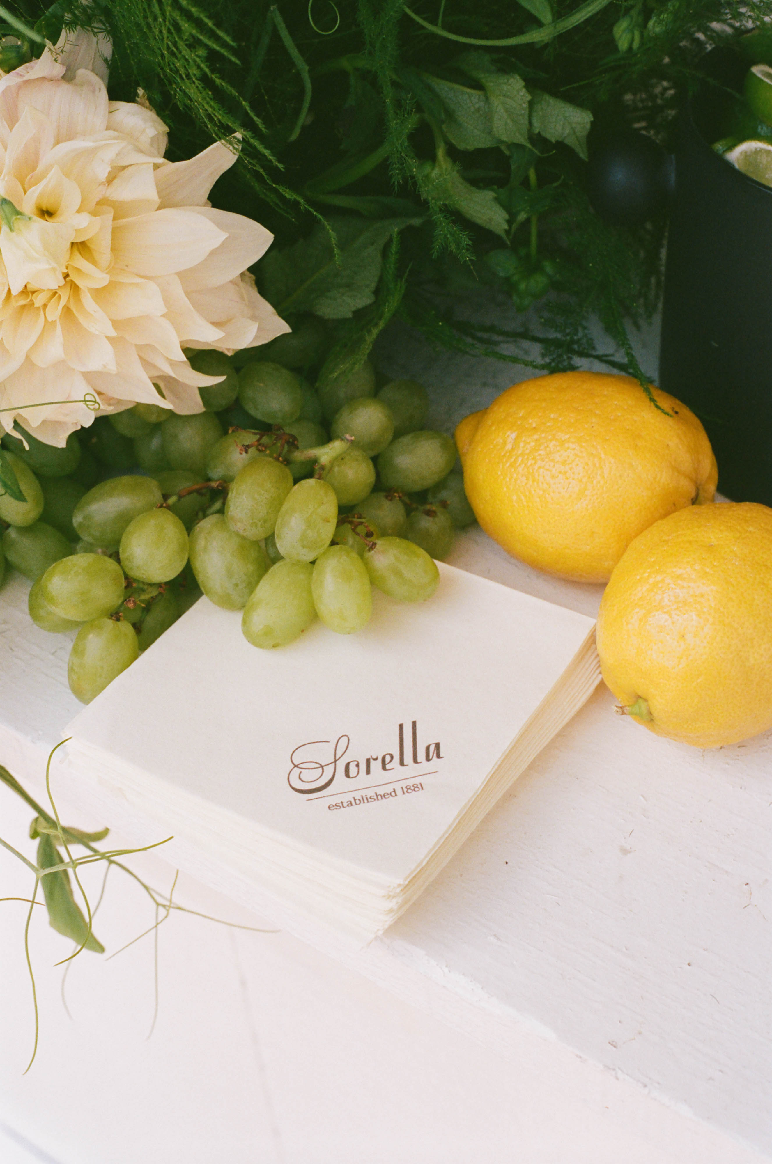 Sorella 1881 branded napkin styled with lemons, grapes, and a soft dahlia bloom — La Dolce Sera detail